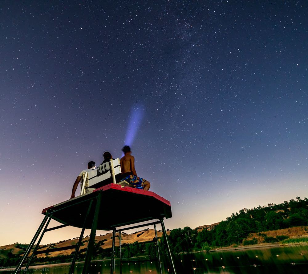 Three boys sit on top of a lifeguard stand and stare out at the dark blue skies covered in shining stars.